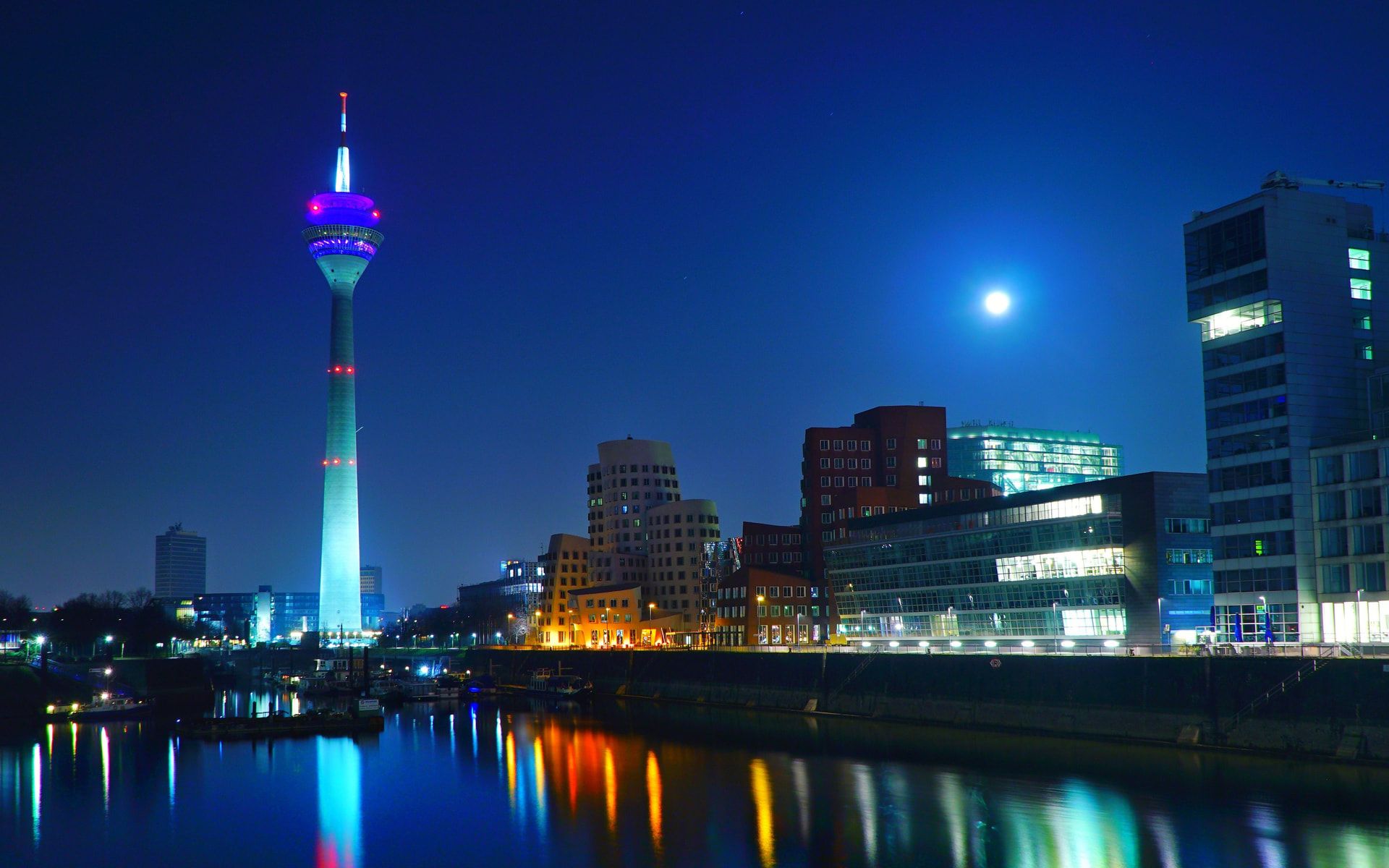 Düsseldorfer Medienhafen bei Nacht: Der beleuchtete Rheinturm und die Hafengebäude spiegeln sich im Rhein, darüber der Mond