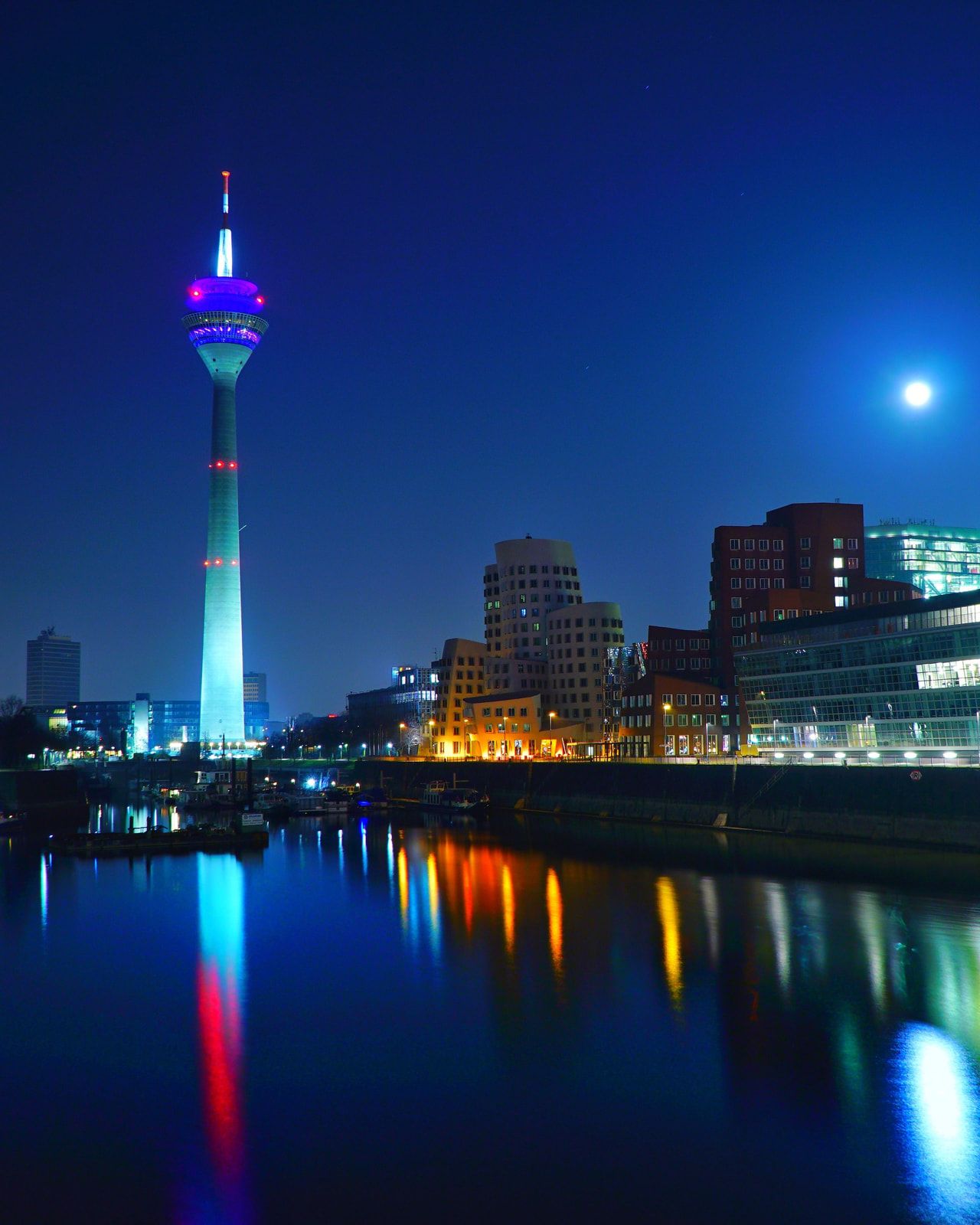 Düsseldorfer Medienhafen bei Nacht: Der beleuchtete Rheinturm und die Hafengebäude spiegeln sich im Rhein, darüber der Mond
